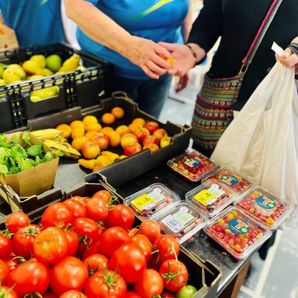 Produce market stall with crates of tomatoes, citrus, bananas and punnets of cherry tomatoes, people browsing nearby