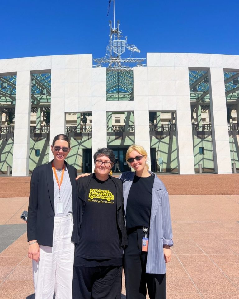 Three women stand smiling outside Australian Parliament House in Canberra under a clear blue sky