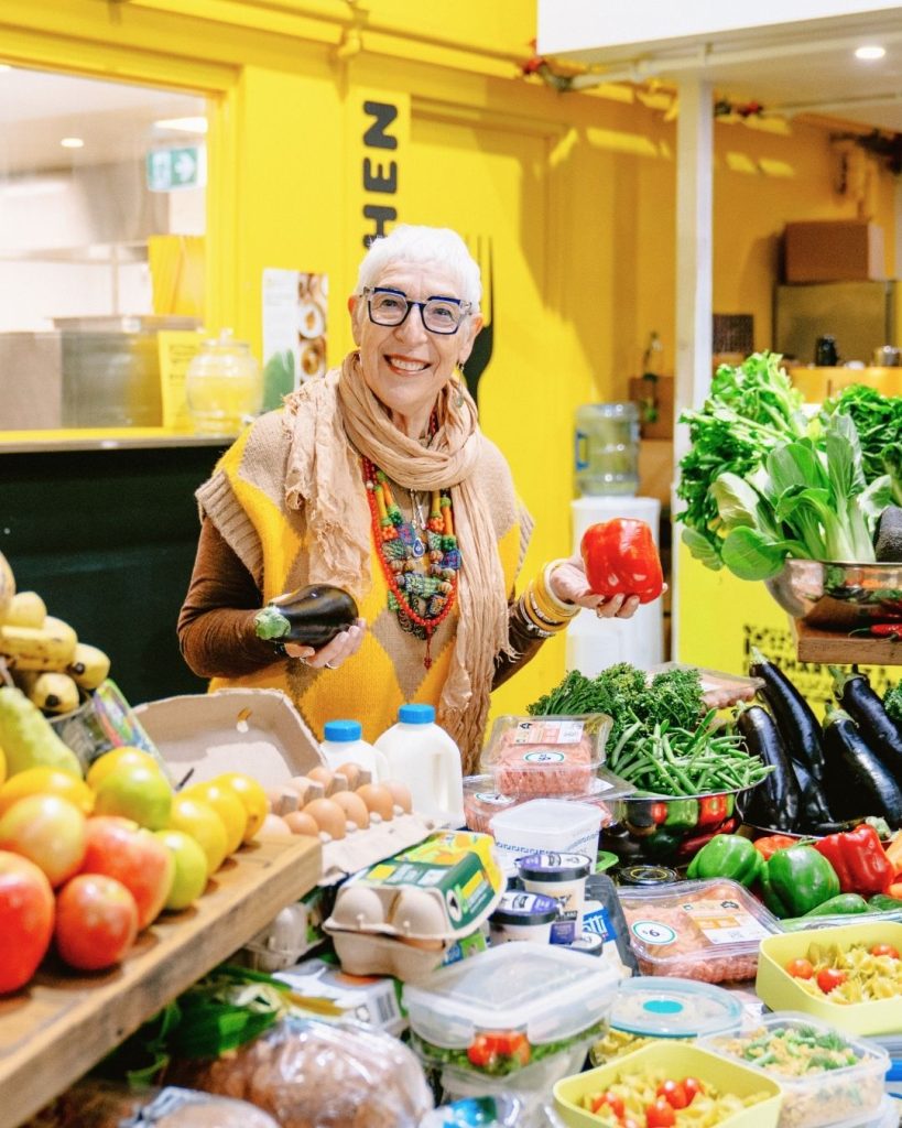 Smiling woman holds an eggplant and red capsicum behind a market table laden with rescued groceries
