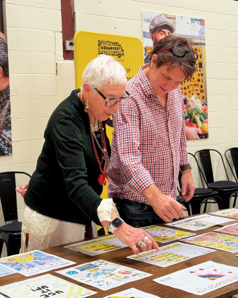 Two people examine colourful 'Use It Up' artwork posters spread across a table at an OzHarvest workshop
