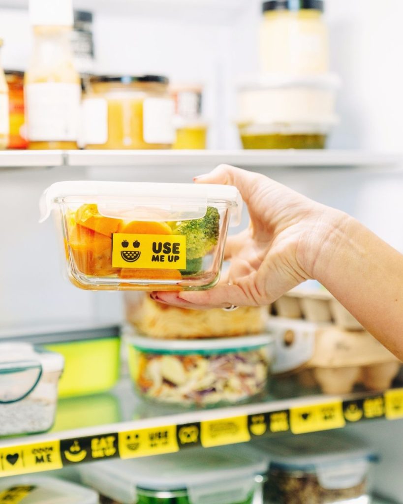 Hand holds a glass container of carrots and broccoli with a yellow 'Use Me Up' label inside a fridge