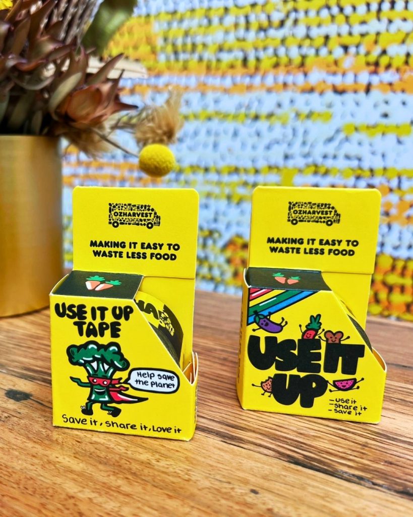 Two yellow OzHarvest Use It Up product boxes displayed on a wooden table against a patterned backdrop