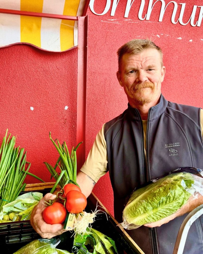 Smiling man in a vest holds ripe tomatoes and lettuce at a market stall against a red community wall