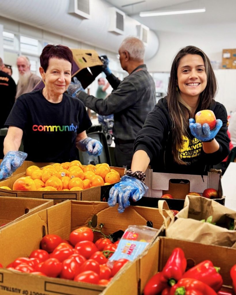 Two smiling volunteers sort rescued oranges, tomatoes and capsicums from boxes at a community food hub