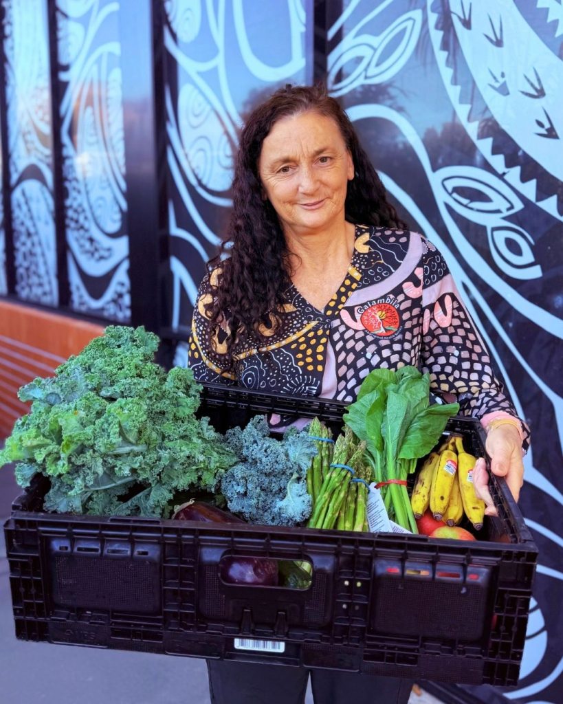 Woman carries a black crate of kale, greens and bananas past a wall with First Nations artwork