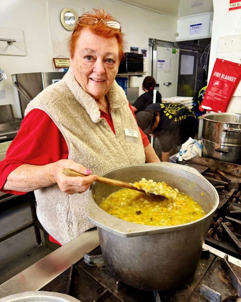 Volunteer in a fleece vest stirs a large pot of soup with a wooden spoon in a community kitchen