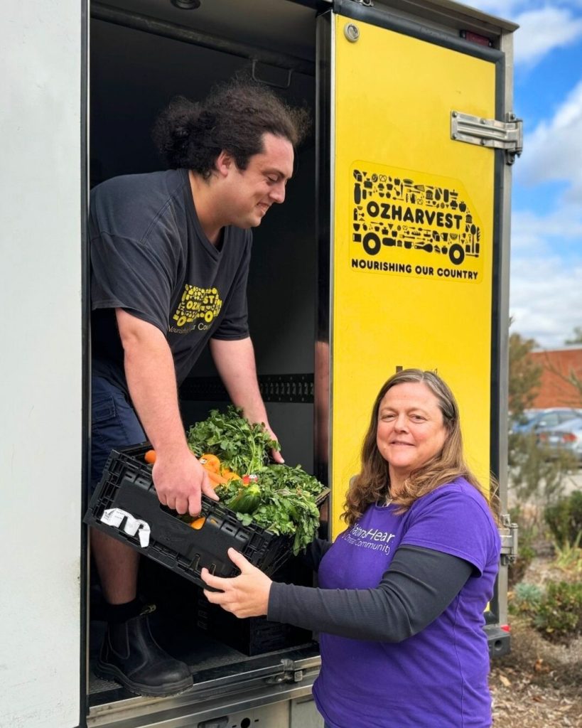 Young man inside a yellow OzHarvest truck hands a crate of vegetables to a smiling woman