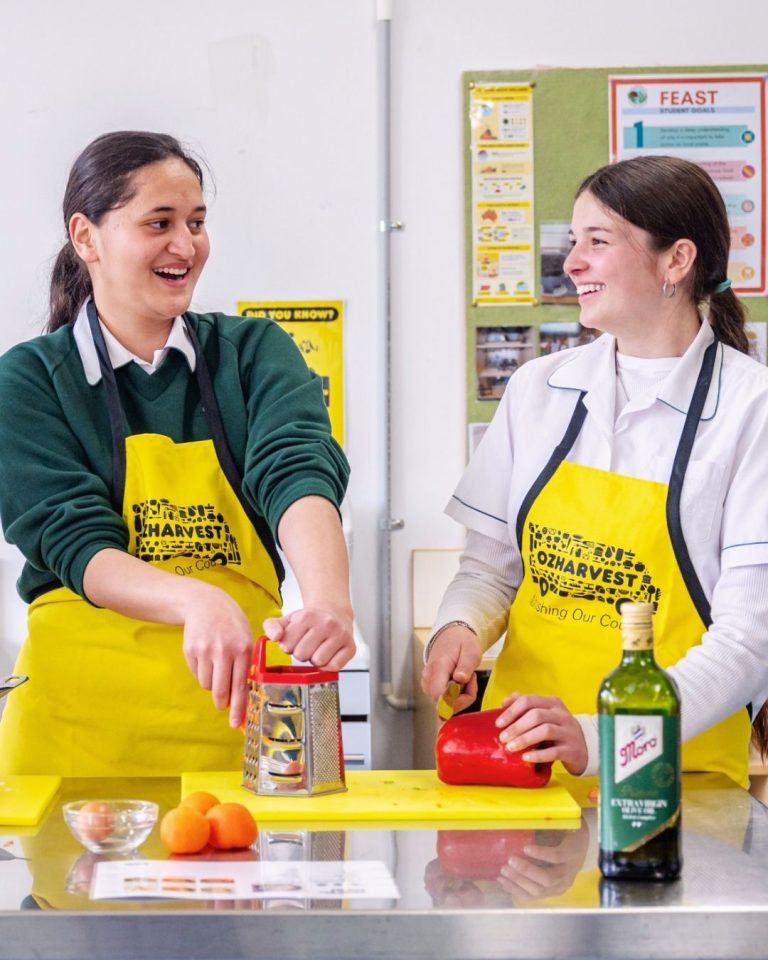 Two students laughing in yellow OzHarvest aprons grate and prepare vegetables in a FEAST classroom kitchen