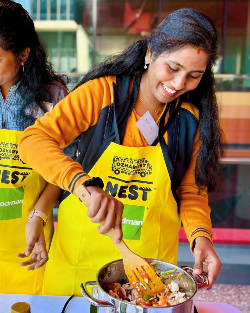 Smiling woman in a yellow OzHarvest NEST apron stirs a pan of stir-fry at an outdoor cooking class