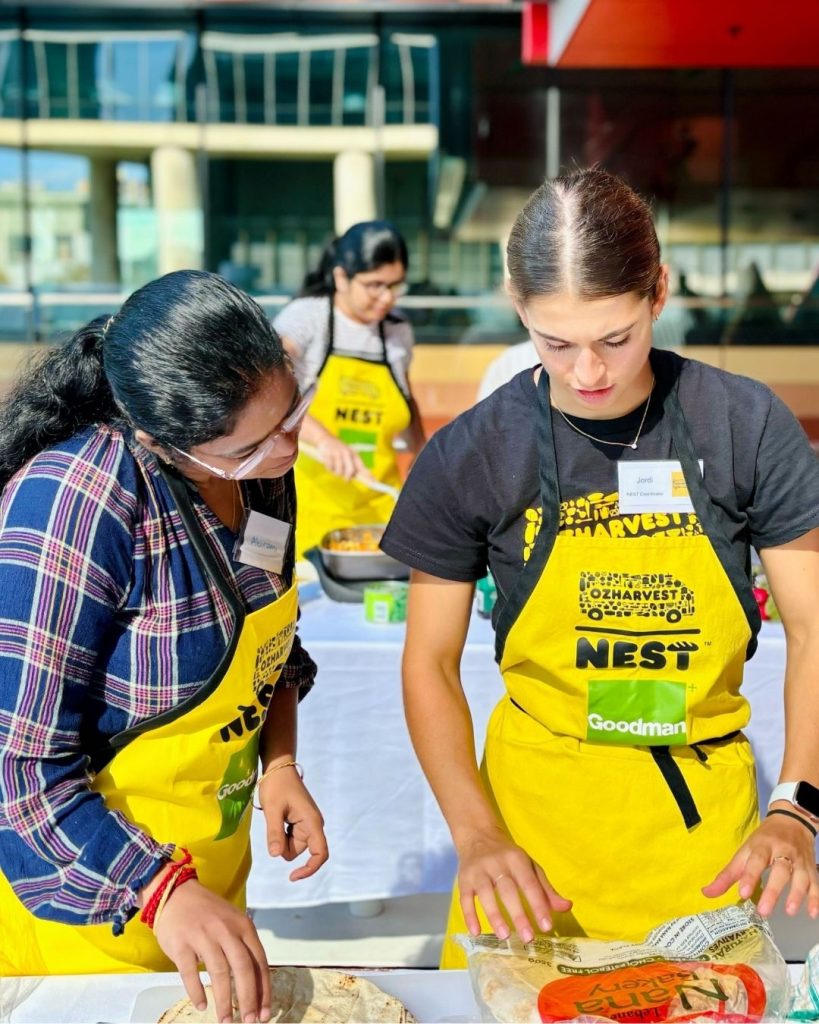 Two women in yellow OzHarvest NEST aprons prepare flatbreads together at an outdoor cooking workshop