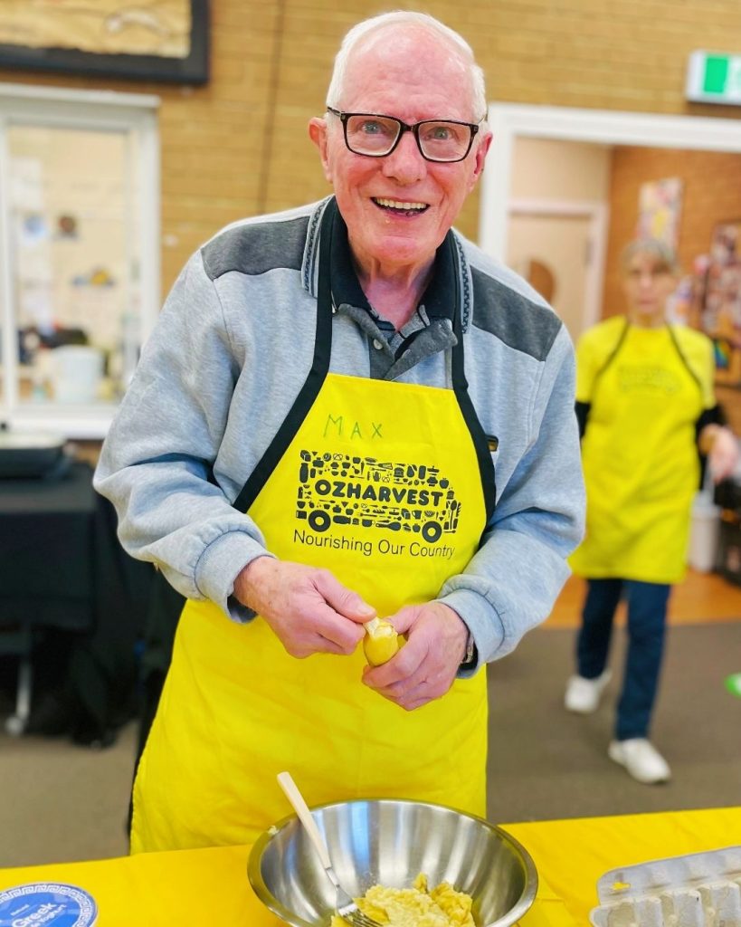 Smiling older man in a yellow OzHarvest apron prepares food at a community kitchen cooking workshop