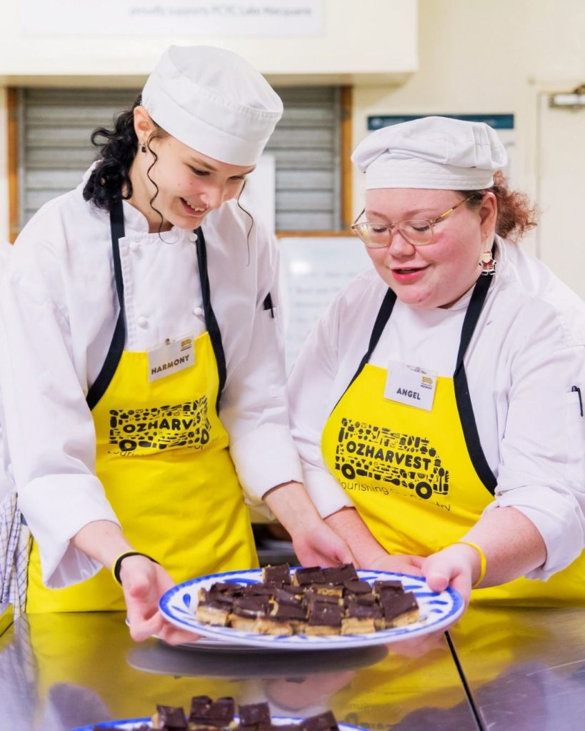 Two OzHarvest Nourish students in chef whites and yellow aprons present a plate of caramel slice