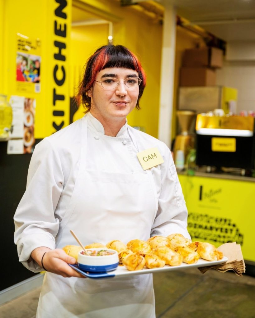 OzHarvest chef holds a tray of golden pastries and salsa in front of a bright yellow Kitchen wall