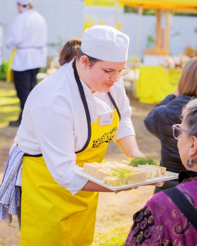 Nourish student chef in yellow apron serves finger sandwiches to a guest at an outdoor OzHarvest event