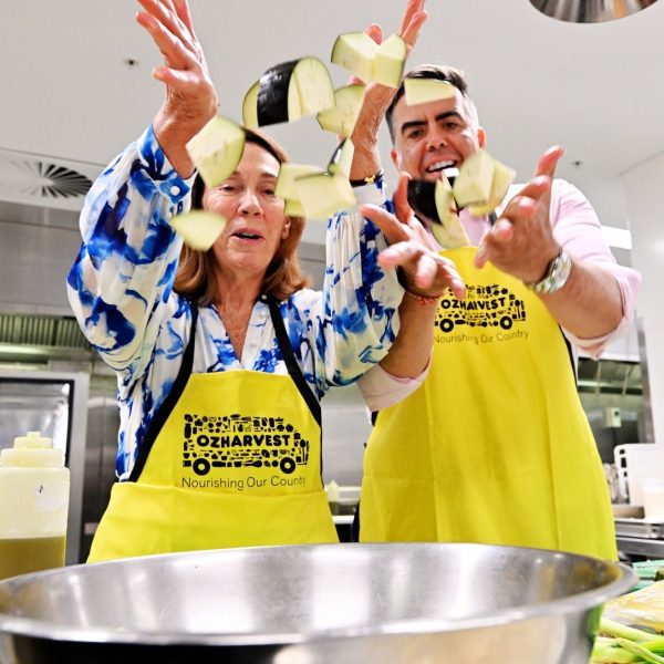 A man and woman in OzHarvest aprons laughing as they toss chopped eggplant above a mixing bowl