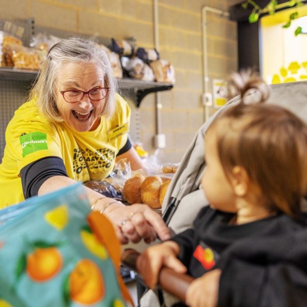 A smiling OzHarvest volunteer greets a toddler in a pram beside shelves of rescued bread