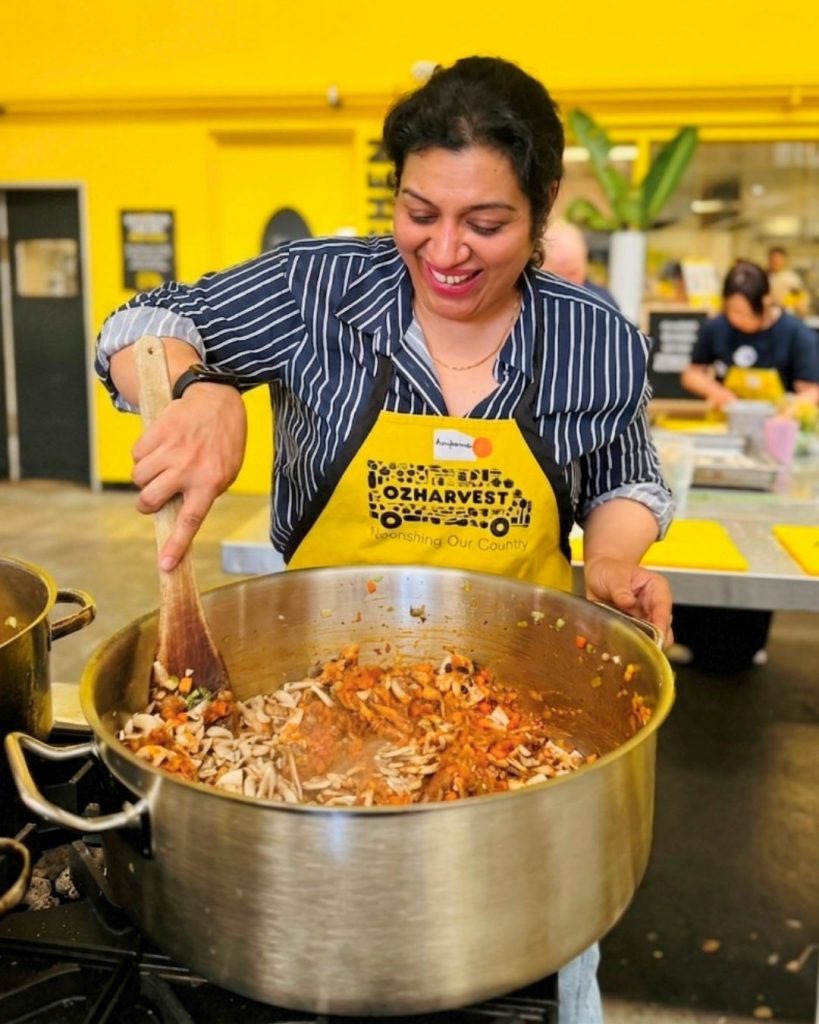 Smiling woman in OzHarvest apron stirring a large pot of vegetables and mushrooms at Cooking for a Cause