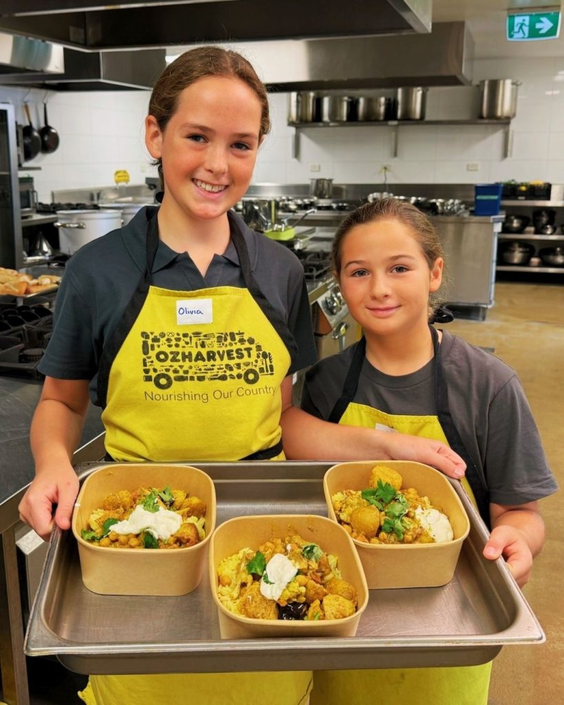 Two girls in OzHarvest aprons holding a tray of cooked meals in a commercial kitchen
