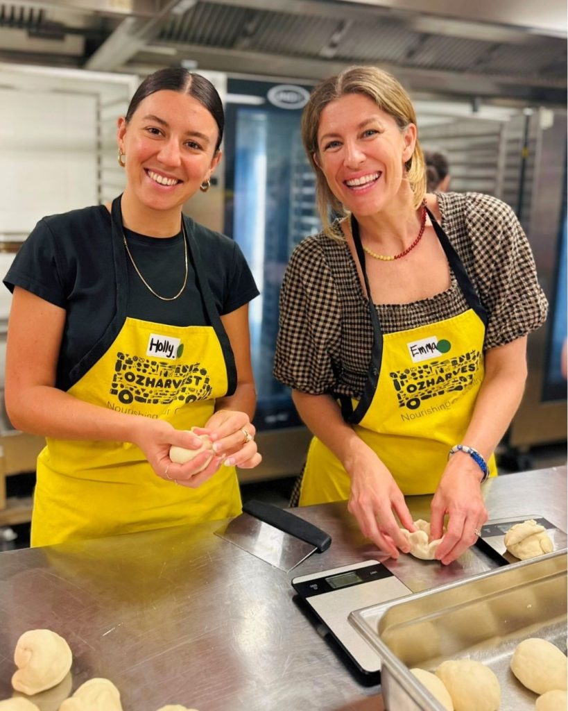 Two smiling women in OzHarvest aprons shaping dough at a Cooking for a Cause kitchen