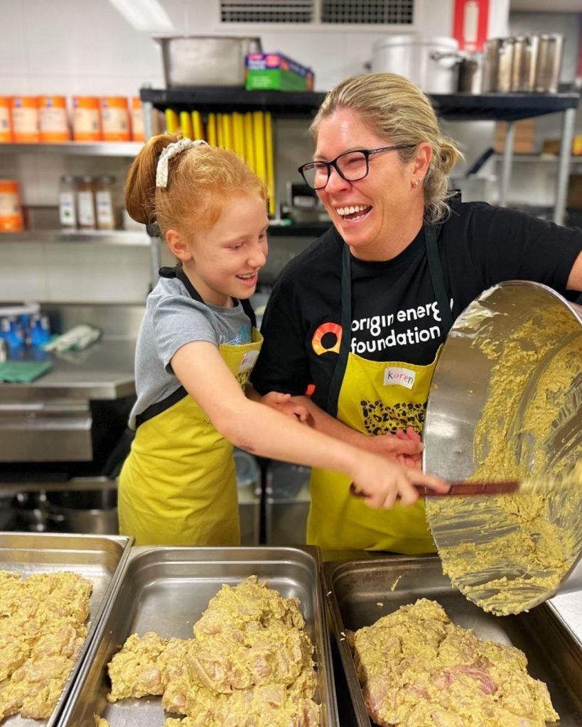 A laughing woman in an Origin Energy Foundation shirt and a girl scoop mixture into kitchen trays