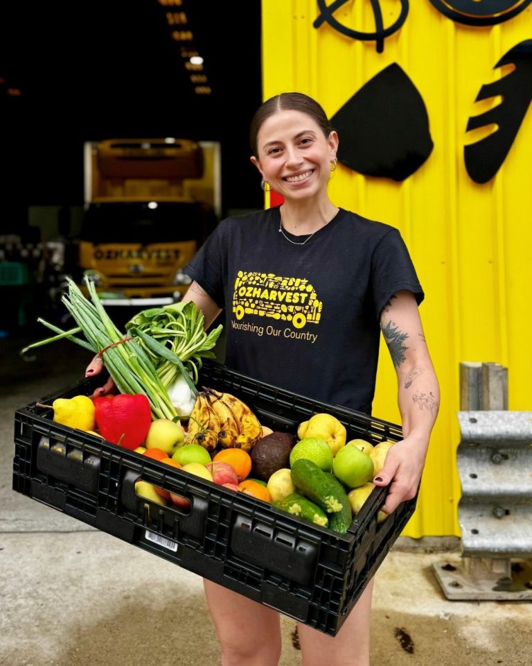 Smiling woman in OzHarvest tee holding a crate of rescued fruit and vegetables by a yellow van