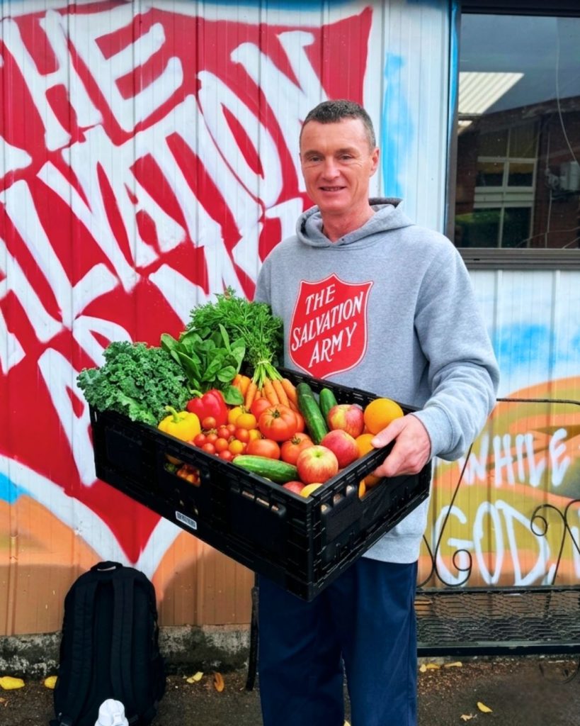 A man in a Salvation Army hoodie holds a crate of colourful fresh vegetables in front of a mural