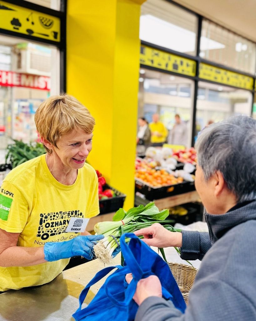 OzHarvest volunteer in yellow shirt hands fresh leafy greens to a shopper at a community market stall