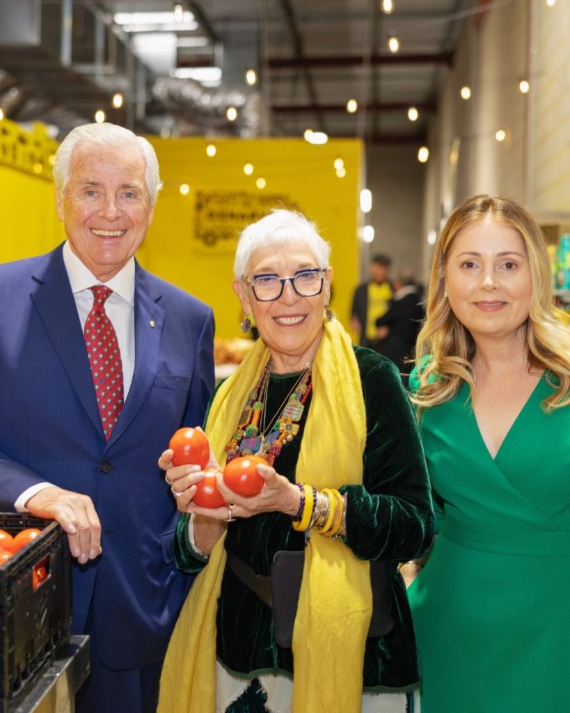 Three well-dressed guests at an OzHarvest warehouse event, the woman in the centre holding fresh tomatoes