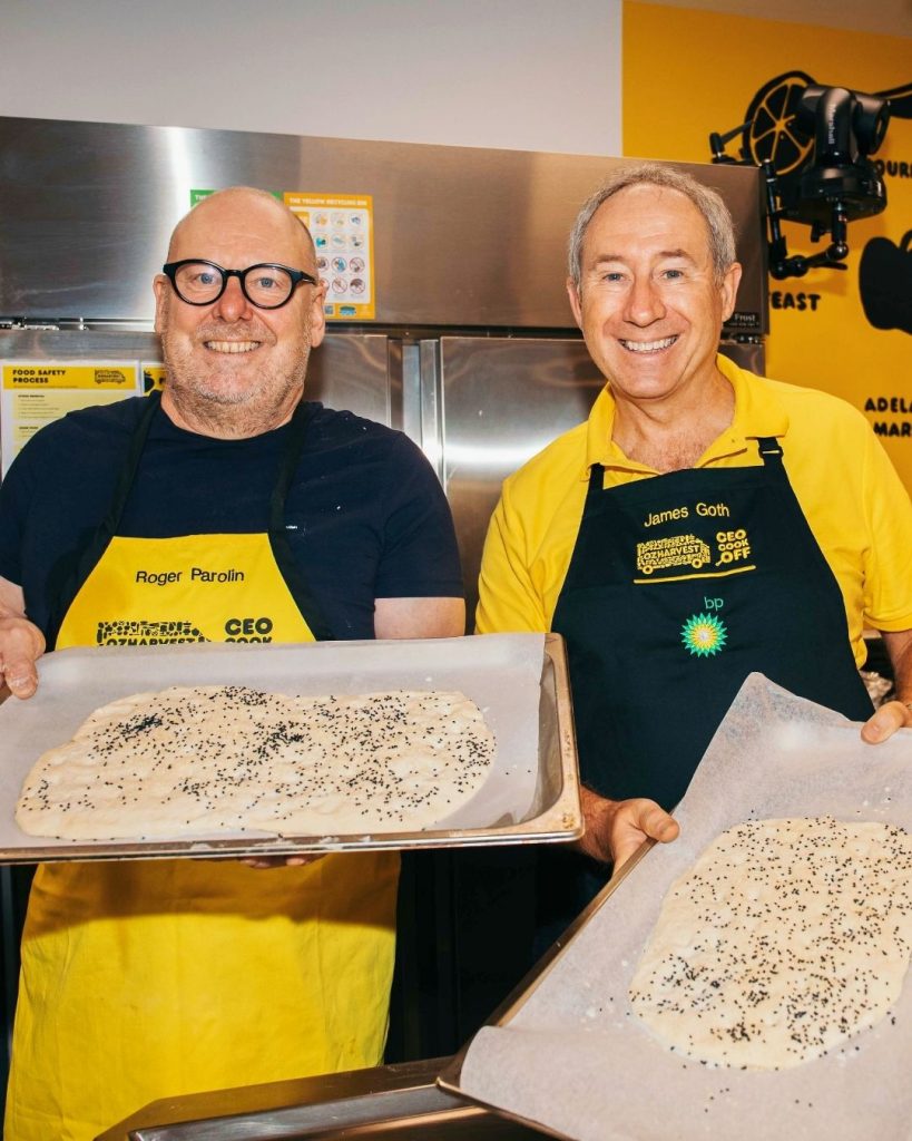 Two men in OzHarvest CEO Cook Off aprons hold baking trays of focaccia in a kitchen