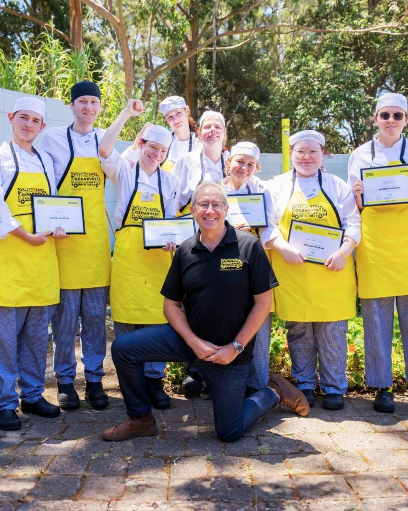 OzHarvest leader kneels with a group of Nourish graduates holding certificates in yellow aprons outdoors