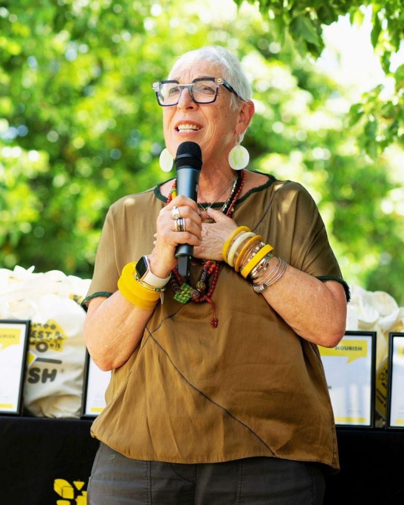 OzHarvest founder speaks into a microphone at an outdoor Nourish event under green trees