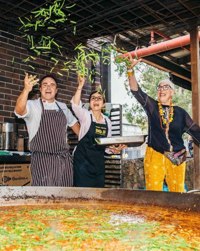 Three chefs including the OzHarvest founder toss green beans over a giant paella pan