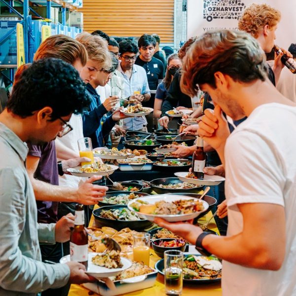 Crowd of people serving themselves from large platters of food along a buffet table at an OzHarvest catering event