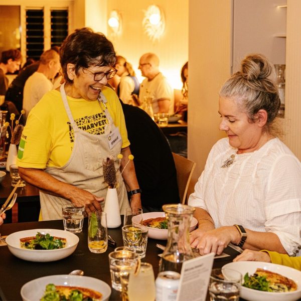 OzHarvest volunteer in yellow shirt chatting and laughing with a seated guest dining at the Refettorio restaurant