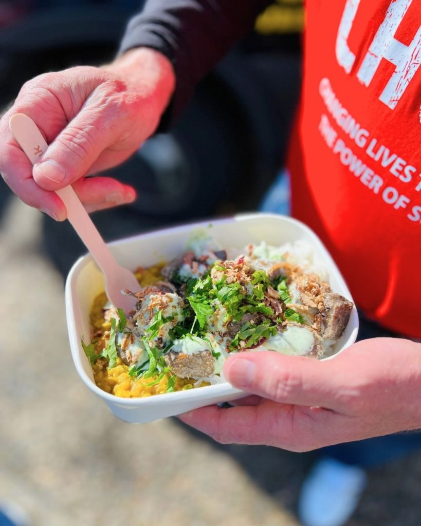 Person in red OzHarvest apron holding takeaway bowl of curry, rice and fresh herbs with wooden fork