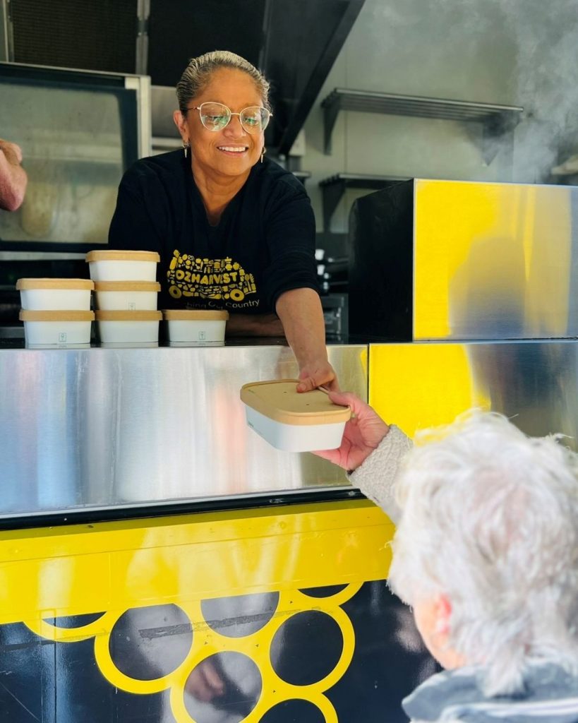Smiling OzHarvest worker hands a takeaway meal container to an older person at a yellow food van window