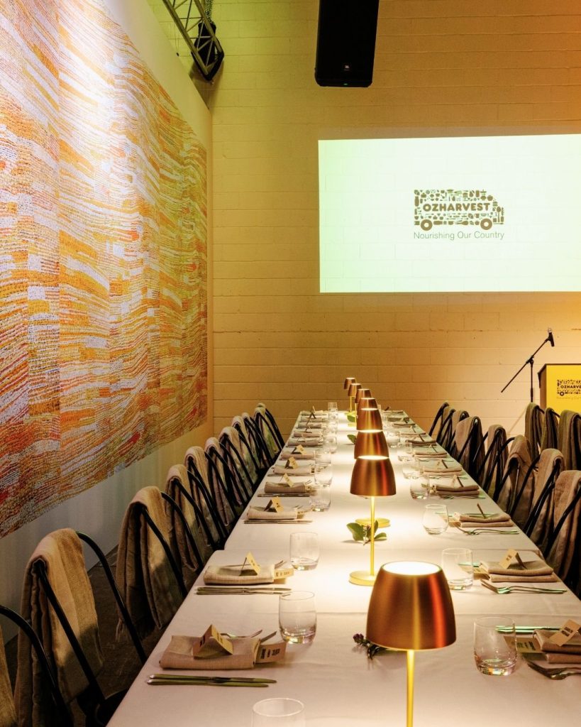 Long banquet table set with brass lamps and an OzHarvest 'Nourishing Our Country' logo projected on wall