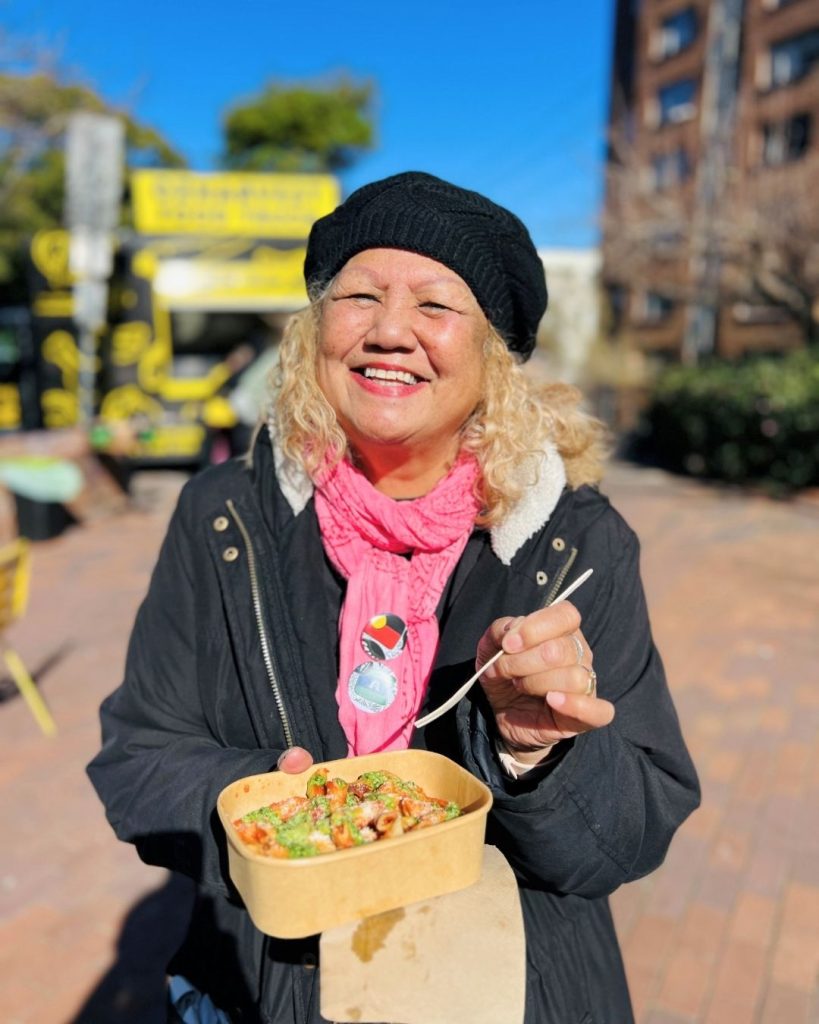 Smiling woman in beret and pink scarf holds a takeaway box of food beside a yellow OzHarvest van