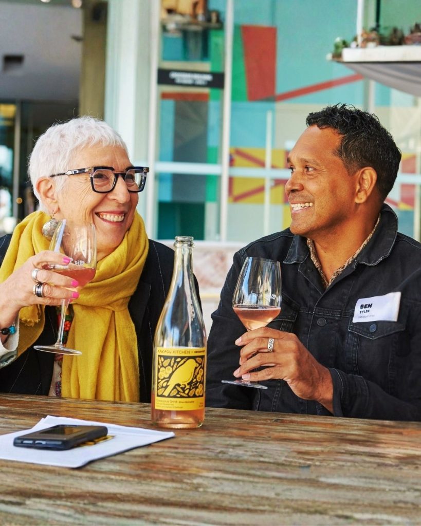 Two people laughing over glasses of rose and a bottle of OzHarvest You Kitchen wine at an outdoor table