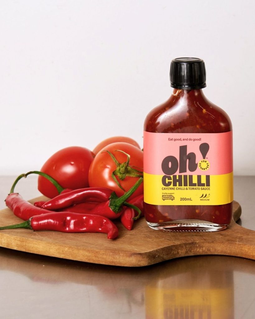 Bottle of Oh! Chilli cayenne and tomato sauce on a board beside fresh red chillies and tomatoes
