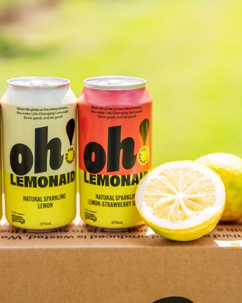 Two cans of Oh! Lemonaid sparkling lemon drinks beside a halved fresh lemon on a kraft box