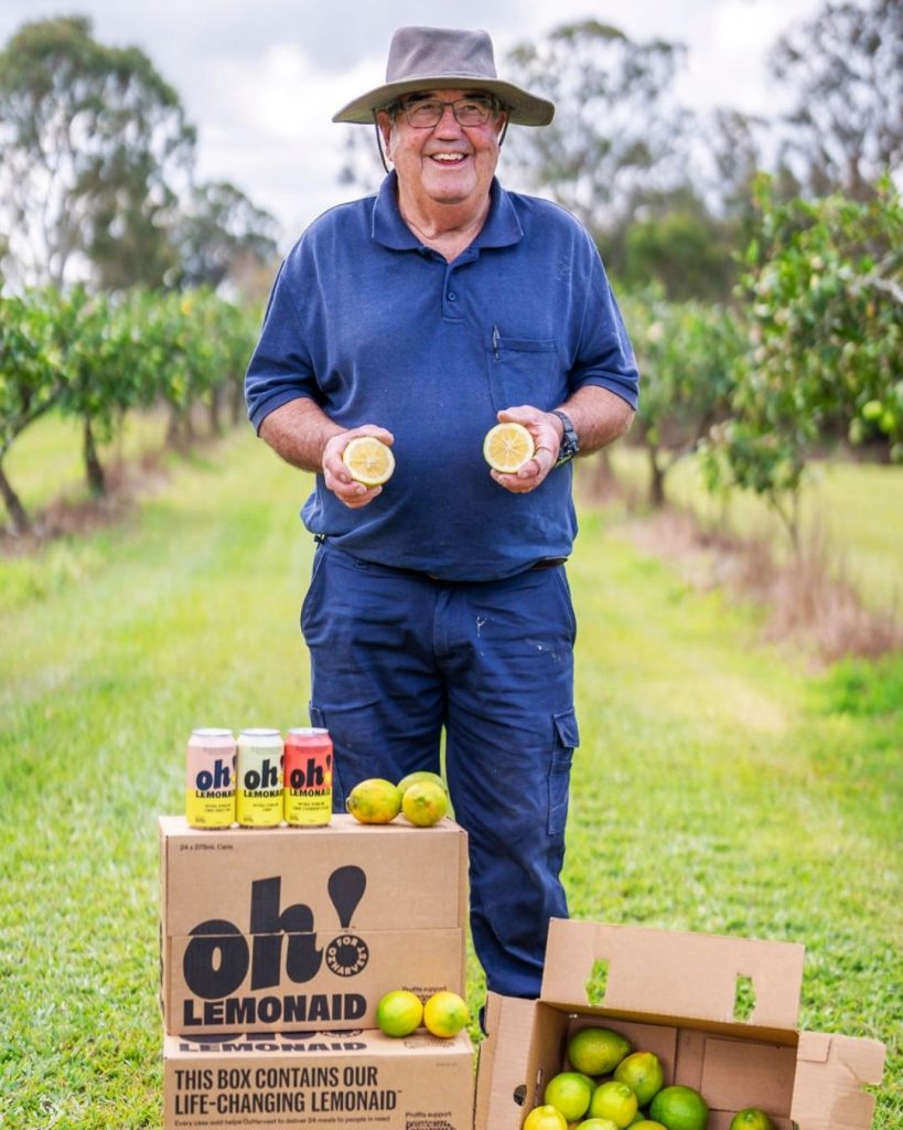 Smiling man in an orchard holds two lemon halves above boxes of Oh! Lemonaid cans and fresh lemons
