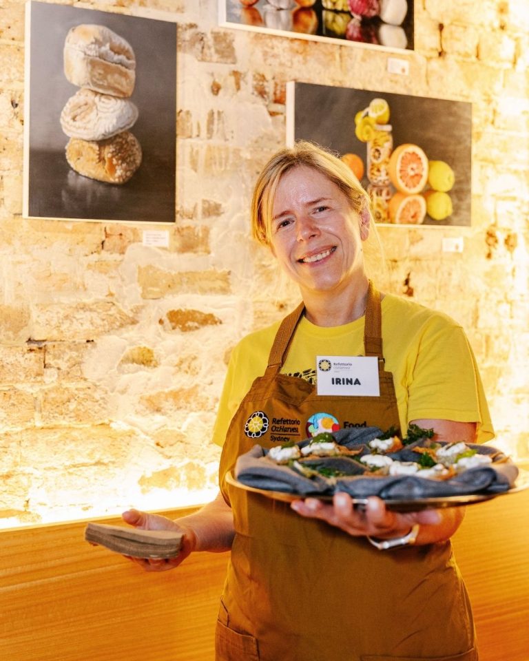 Refettorio volunteer Irina in a yellow shirt holds a plate of canapes by framed food photos on a stone wall
