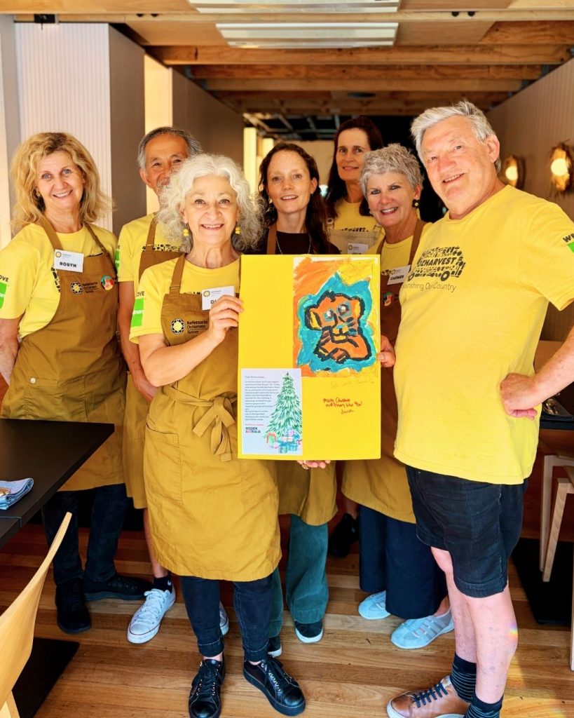 Group of OzHarvest volunteers in yellow aprons and shirts holding a child's painted artwork in a restaurant