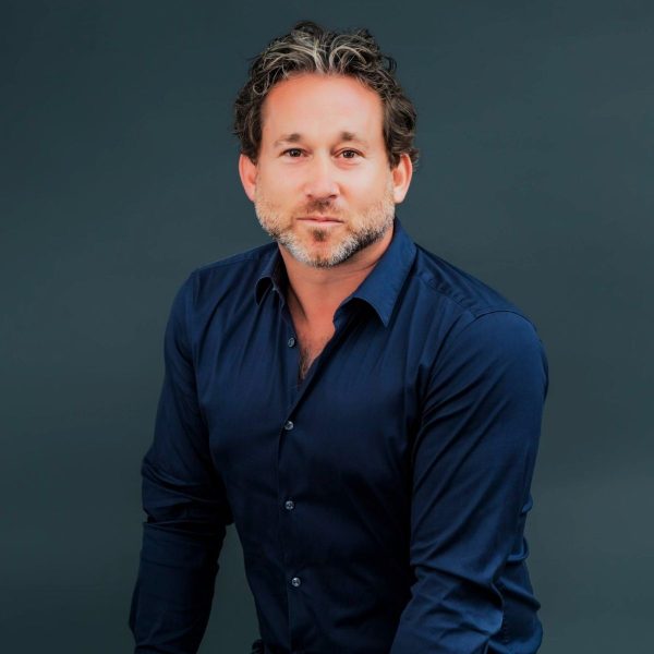 Man with curly greying hair and a short beard wearing a navy shirt, against a dark teal backdrop