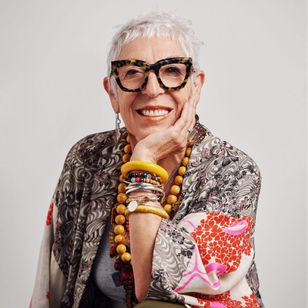 Smiling older woman with white hair, bold glasses, yellow bangles and a patterned scarf, hand to cheek