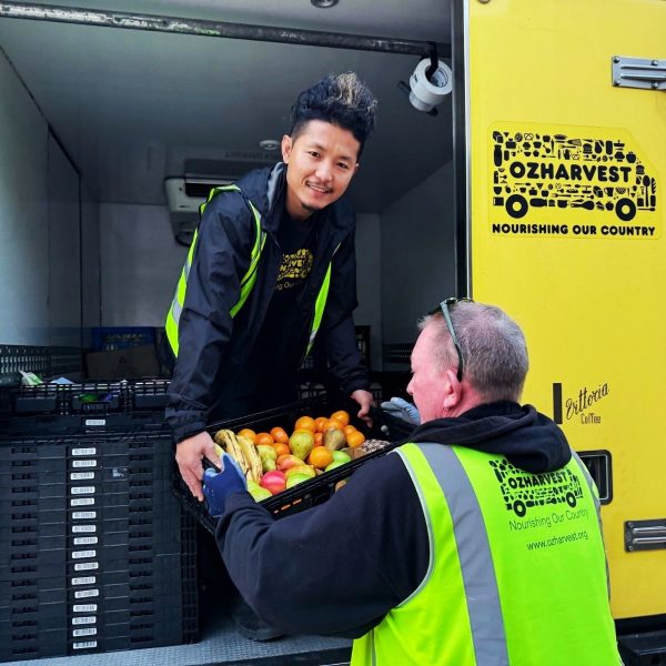 Two OzHarvest workers in hi-vis vests passing a crate of rescued fruit at the back of a yellow van