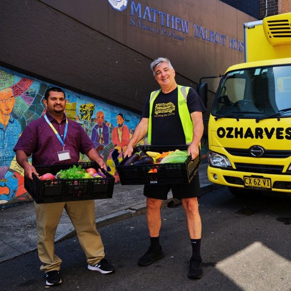 Two OzHarvest workers carry crates of fresh vegetables beside a yellow truck outside Matthew Talbot Hostel