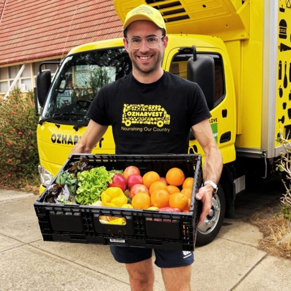 Man in OzHarvest cap and shirt holds a crate of tomatoes, capsicums and lettuce in front of a yellow truck