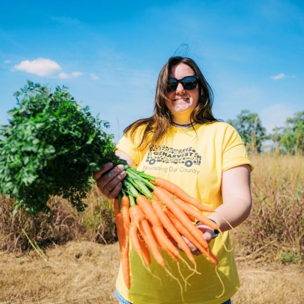 Woman in yellow OzHarvest t-shirt and sunglasses holds a bunch of fresh carrots in a sunlit field